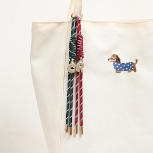 Könnte beinhalten: Cremefarbene Tragetasche mit einer Dackel-Stickerei, die einen blauen Mantel mit weißen Punkten trägt. Die Tasche hat grün-rot gestreifte Kordeln mit goldfarbenen Metallspitzen und Charms mit der Aufschrift "CR".