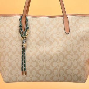 May include: Beige tote bag with a repeating pattern and brown leather handles. A green and white striped charm with a gold star and the name "JENNY" hangs from the handle. The bag has a light brown trim.