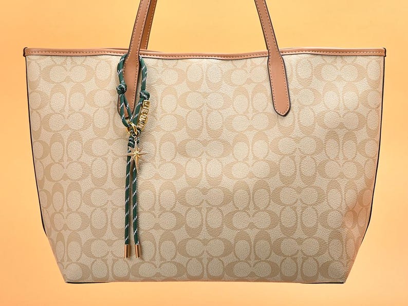 May include: Beige tote bag with a repeating pattern and brown leather handles. A green and white striped charm with a gold star and the name "JENNY" hangs from the handle. The bag has a light brown trim.
