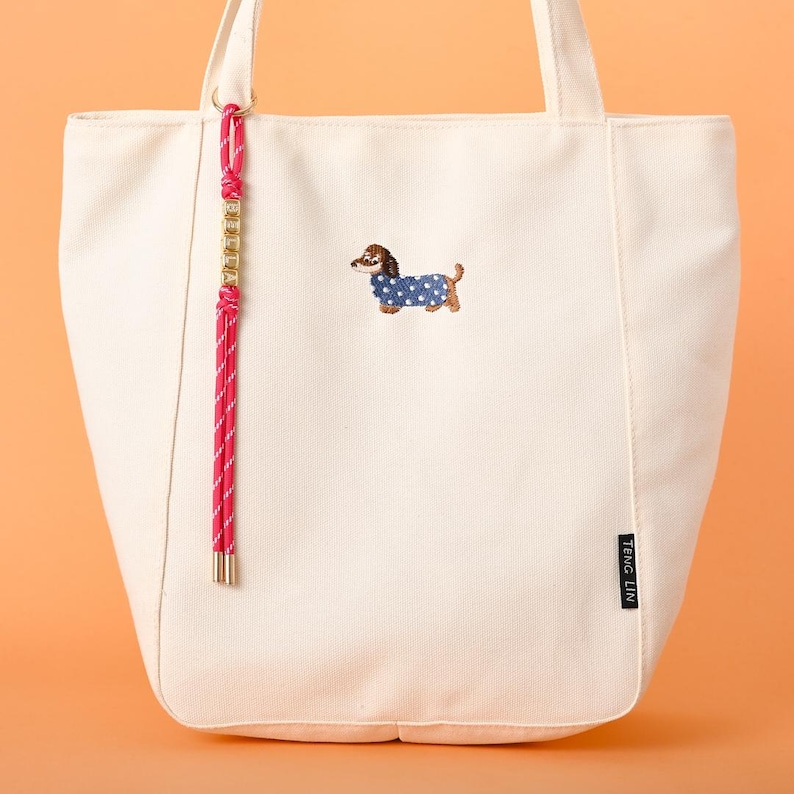 May include: Cream-colored tote bag with a dachshund dog embroidery wearing a blue polka-dot coat. The bag has a pink and white rope handle and a gold-colored chain detail. The brand name "TENG LIN" is printed on a black label.