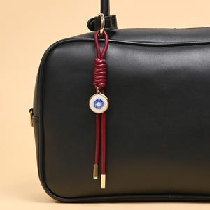 Può includere: Una borsa in pelle nera con un ciondolo decorativo attaccato al manico. Il ciondolo è composto da un cordino annodato rosso scuro, un pendente circolare con un motivo a occhio blu e bianco e due cordini pendenti con punte in metallo dorato.