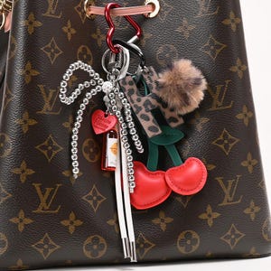 Puede incluir: Un bolso marrón con estampado de monograma, adornado con una colección de abalorios. Los abalorios incluyen un corazón rojo, una botella en miniatura, una letra "H" con estampado de leopardo, un pompón esponjoso y un par de cerezas de cuero rojo.