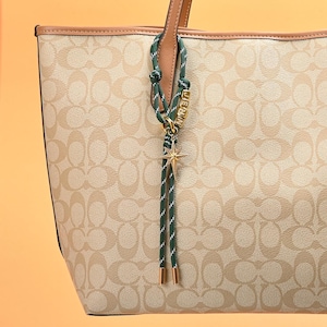 Może przedstawiać: Beżowa torba typu tote z powtarzającym się wzorem logo. Torba ma brązowe skórzane uchwyty i wykończenia. Zielony i biały sznurkowy urok z napisem "JENNA" i złotym urokiem w kształcie gwiazdy wisi z uchwytu.