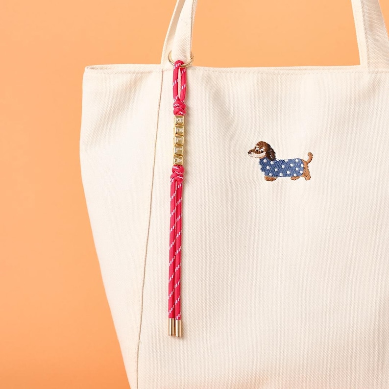 May include: Cream-colored tote bag with a pink and white rope handle and a gold-colored chain detail. The bag features an embroidered dachshund wearing a blue coat with white polka dots. The bag has two handles.