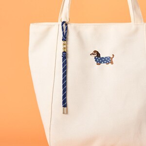 May include: Cream-colored tote bag with a blue and gold accent. The bag features an embroidered dachshund wearing a blue polka-dot sweater. A navy blue and gold rope charm hangs from the handle. The bag is made of a sturdy canvas material.