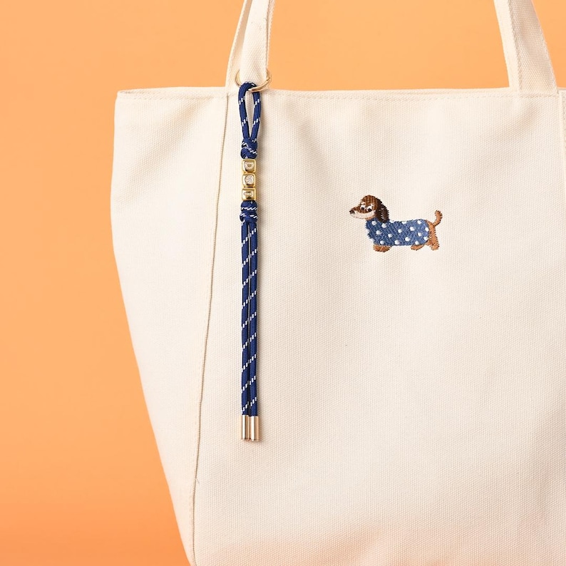 May include: Cream-colored tote bag with a blue and gold accent. The bag features an embroidered dachshund wearing a blue polka-dot sweater. A navy blue and gold rope charm hangs from the handle. The bag is made of a sturdy canvas material.