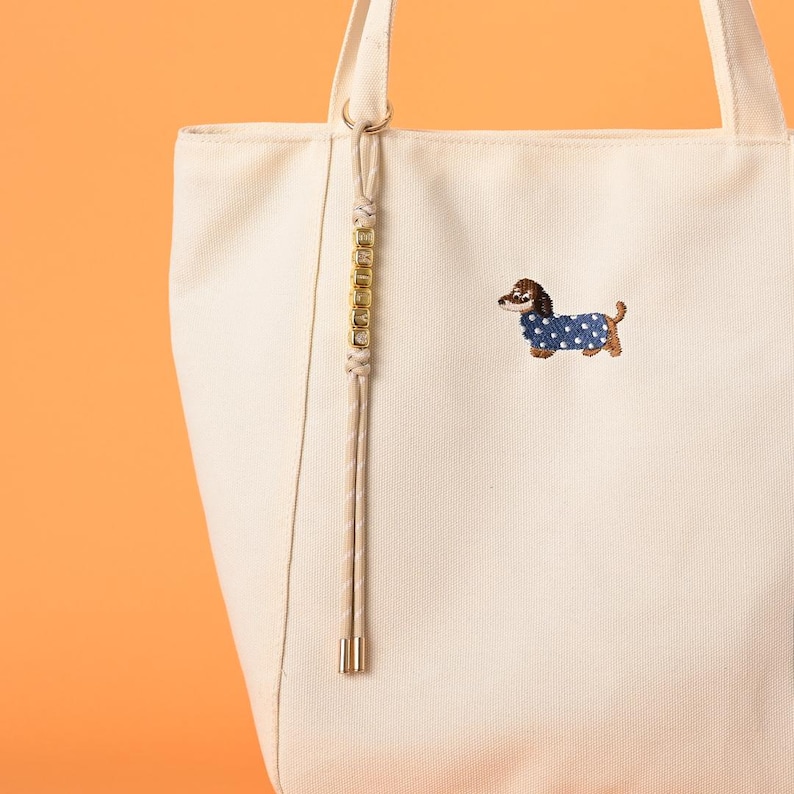 May include: Cream-colored tote bag with a brown and blue embroidered dachshund wearing a polka-dot coat. The bag has a long, decorative strap with gold-colored accents and a cream-colored handle.