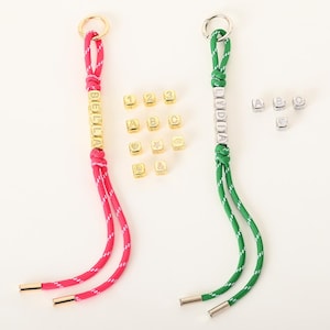 May include: Two keychains with personalized names. One keychain is pink and white, with the name "BELLA" in gold letters. The other is green and white, with the name "LYDIA" in silver letters. Additional gold and silver letter beads are scattered around.