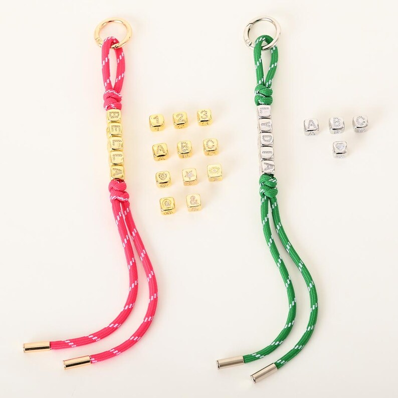 May include: Two keychains with personalized names. One keychain is pink and white, with the name "BELLA" in gold letters. The other is green and white, with the name "LYDIA" in silver letters. Additional gold and silver letter beads are scattered around.