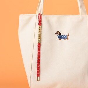 May include: Off-white canvas tote bag with a red and white striped rope detail and gold-colored accents. The bag features an embroidered dachshund wearing a blue polka-dot sweater. The word "CAROLINE" is spelled out in gold letters.