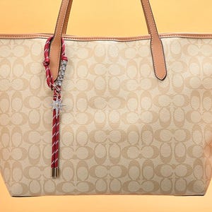 May include: Beige tote bag with a repeating pattern and brown leather handles. A red and white striped charm with a star-shaped pendant hangs from the handle. The bag has a structured shape and a light-colored interior.