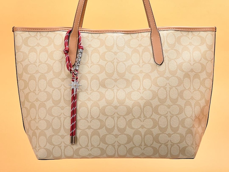 May include: Beige tote bag with a repeating pattern and brown leather handles. A red and white striped charm with a star-shaped pendant hangs from the handle. The bag has a structured shape and a light-colored interior.