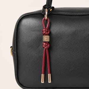Puede incluir: Un bolso de mano de cuero negro con un colgante de cordón rojo oscuro atado en dos nudos. Una cuenta cuadrada de tono dorado con la letra E grabada en su cara frontal está colocada entre los nudos. El colgante cuelga de una anilla de metal dorado.