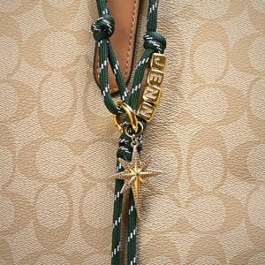 May include: A green and white rope charm with gold-colored hardware and a star pendant. The charm spells out the name "JENNA" in gold letter beads. The background is a tan and brown pattern.
