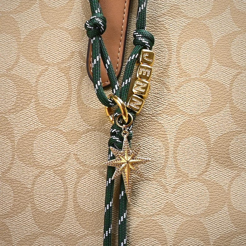 May include: A green and white rope charm with gold-colored hardware and a star pendant. The charm spells out the name "JENNA" in gold letter beads. The background is a tan and brown pattern.
