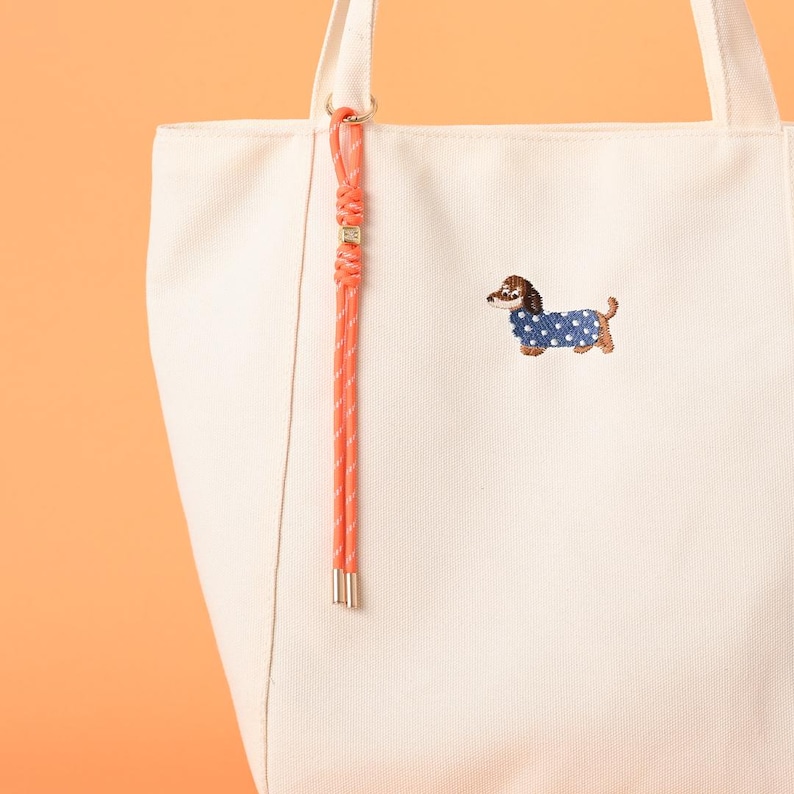 May include: Cream-colored canvas tote bag with an embroidered dachshund wearing a blue polka-dot sweater. The bag has a coral-colored rope detail and a matching accent on the handle. The bag is a stylish accessory.