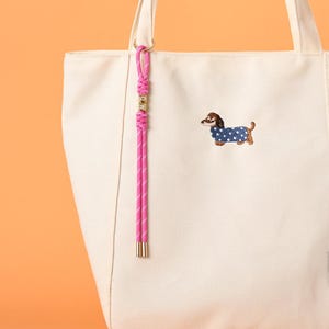 May include: Cream-colored tote bag with a pink and white striped lanyard and a small embroidered dachshund wearing a blue polka-dot coat. The lanyard has gold-colored metal accents and a metal end.