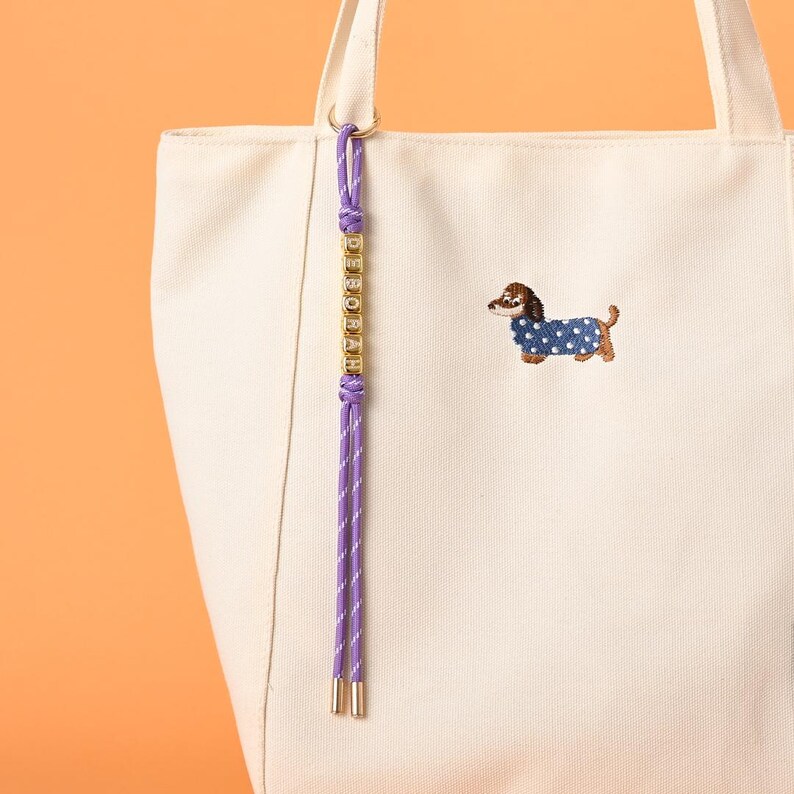 May include: Cream-colored tote bag with a purple and white cord detail and gold-colored accents. The bag features an embroidered dachshund wearing a blue polka-dot coat. A charm with the name "DEBORAH" hangs from the bag.
