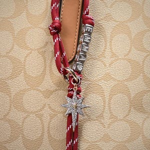 May include: Close-up of a red and white rope bag charm with a silver starburst pendant. The charm includes silver lettered cubes spelling "EMMA". The background is a light brown pattern.