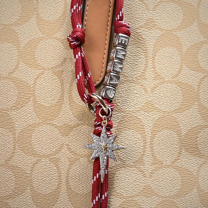 May include: Close-up of a red and white rope bag charm with a silver starburst pendant. The charm includes silver lettered cubes spelling "EMMA". The background is a light brown pattern.
