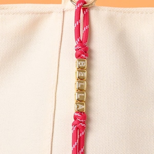 May include: A close-up of a pink and white rope keychain with gold letter beads spelling "DELILA". The keychain is attached to a beige canvas tote bag with a gold ring. The rope is knotted at the top and bottom of the beads.