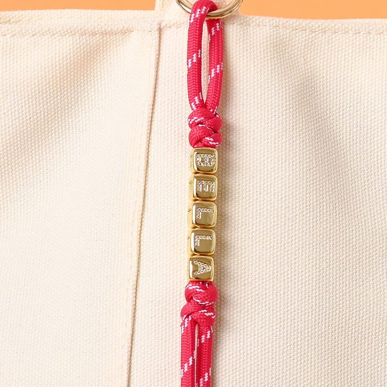 May include: A close-up of a pink and white rope keychain with gold letter beads spelling "DELILA". The keychain is attached to a beige canvas tote bag with a gold ring. The rope is knotted at the top and bottom of the beads.