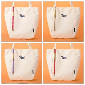 May include: Four off-white tote bags with a dachshund dog embroidered on the front. Each bag has a different colored decorative cord attached to the handle: pink, beige, purple, and red. The bags are on an orange background.