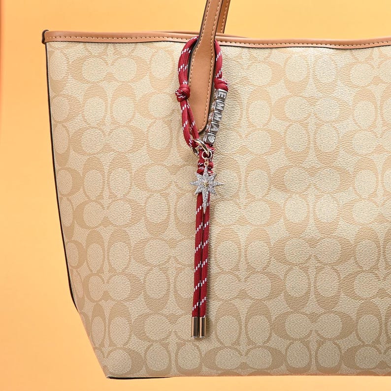May include: Beige tote bag with a repeating pattern. A red and white striped charm with the name "EMMA" and a starburst charm hangs from the handle. The bag has a light brown trim.