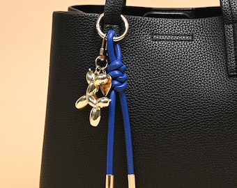 Könnte beinhalten: Eine schwarze Tragetasche aus strukturiertem Leder mit einem silberfarbenen Metallring. Am Ring hängen ein goldfarbener Ballonhund-Anhänger und eine blaue Lederkordel, die oben verknotet ist und unten zwei silberfarbene Metallkappen aufweist.