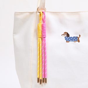 May include: A cream-coloured canvas tote bag featuring an embroidered brown dachshund wearing a blue polka-dot sweater. Attached to the bag's handle is a decorative accessory consisting of two knotted cords in bright yellow and hot pink colours.