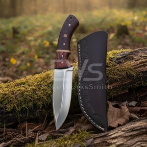Könnte beinhalten: Ein handgefertigtes Messer mit einem dunklen Holzgriff und einer polierten Stahlklinge. Das Messer wird mit einer schwarzen Lederscheide geliefert. Der Griff hat Messingakzente und dekorative Stifte. Messer und Scheide werden auf einem moosbedeckten Baumstamm präsentiert.