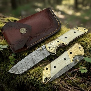 Puede incluir: Un cuchillo plegable con una hoja estampada y un mango color crema con detalles dorados. El cuchillo est&aacute; abierto, revelando la hoja, y est&aacute; junto a un cuchillo cerrado y una funda de cuero marr&oacute;n. El cuchillo est&aacute; sobre una superficie musgosa.