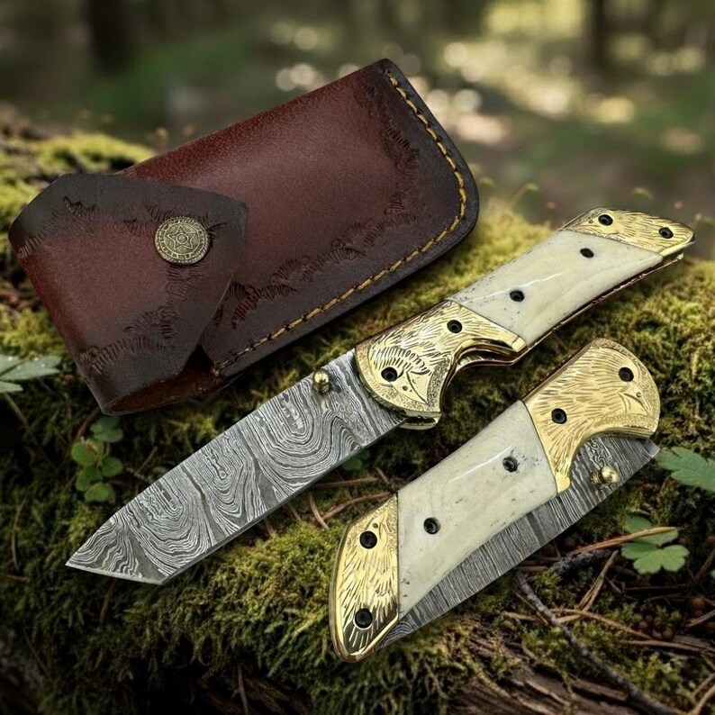 Puede incluir: Un cuchillo plegable con una hoja estampada y un mango color crema con detalles dorados. El cuchillo est&aacute; abierto, revelando la hoja, y est&aacute; junto a un cuchillo cerrado y una funda de cuero marr&oacute;n. El cuchillo est&aacute; sobre una superficie musgosa.