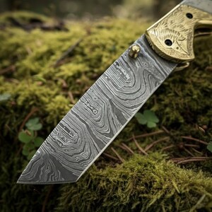 Puede incluir: Primer plano de una hoja de cuchillo plegable de acero de Damasco con un patr&oacute;n detallado de vetas de madera. El mango es de color dorado con un dise&ntilde;o grabado. El cuchillo descansa sobre musgo verde.