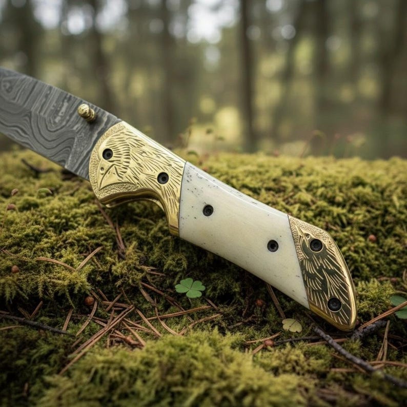 Puede incluir: Primer plano de un cuchillo de acero de Damasco con mango crema y detalles dorados con forma de cabeza de &aacute;guila. El cuchillo descansa sobre musgo verde, mostrando su intrincado dise&ntilde;o y artesan&iacute;a.