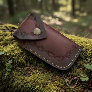 Puede incluir: Una funda de cuchillo de cuero marr&oacute;n con relieve floral y costuras amarillas. La funda tiene una solapa asegurada por un bot&oacute;n de metal con un dise&ntilde;o circular. El fondo es un suelo forestal cubierto de musgo.