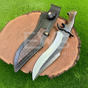 May include: A large hunting knife with a polished steel blade and a textured, dark handle. The knife is paired with a black leather sheath with a brown snakeskin-like accent. The knife and sheath rest on a wooden surface.