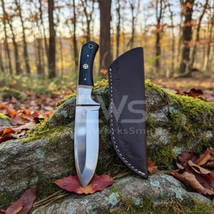 Puede incluir: Un cuchillo de caza artesanal con una hoja de acero pulido y un mango negro con detalles en latón. El cuchillo se muestra con una funda de cuero marrón, sobre un fondo de roca natural y musgosa.