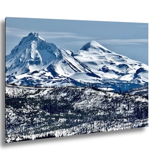 Op de afbeelding: Een landschapsfoto met twee met sneeuw bedekte bergtoppen onder een heldere blauwe lucht. De bergen zijn het middelpunt, met een voorgrond van donkere bomen en een hint van een weg. De afbeelding heeft een koel kleurenpalet.