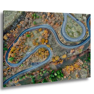Puede incluir: Vista aérea de una carretera de montaña sinuosa con curvas cerradas. La carretera está rodeada de árboles con follaje otoñal en tonos naranja, amarillo y verde. Varios coches son visibles en la carretera y el cielo está despejado.