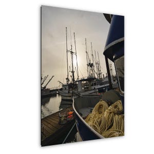 Puede incluir: Fotografía de un puerto con barcos de pesca atracados en un muelle de madera. Los barcos tienen mástiles altos y están silueteados contra el sol. Un barco azul en primer plano tiene cuerdas amarillas enrolladas.