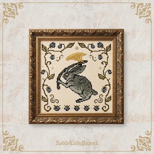 Peut inclure: Une broderie au point de croix représentant un lapin gris tenant une hache dorée, encadrée dans un cadre doré orné. Le motif est entouré d'une bordure de baies bleu foncé et de feuilles vertes sur un fond en tissu couleur crème.