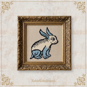 May include: A cross-stitch embroidery of a rabbit with blue ears and blue legs on a beige fabric background, enclosed in an ornate, antique-style gold frame. The rabbit is depicted in profile, facing right, with black outlines and blue accents.