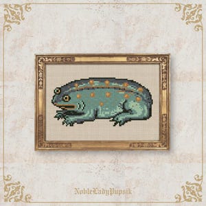 Puede incluir: Arte de punto de cruz de una rana en un marco dorado. La rana es de color turquesa con manchas naranjas y una espalda azul oscuro. El marco tiene detalles ornamentados. El texto "NobleLadyPupsik" está en la parte inferior.
