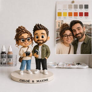 May include: A custom figurine of a couple, with the names "CHLOÉ & MAXIME" on the base. The figures are wearing glasses and casual clothing. The woman holds a camera, and the man holds a book. A photo of the couple is in the background.