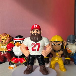 May include: A group of colourful figurines, including a muscular figure wearing a white t-shirt with the number "27" and a red cap. Other figures include a lion, penguin, monkey, and shark. The figures are displayed on a white rotating platform.