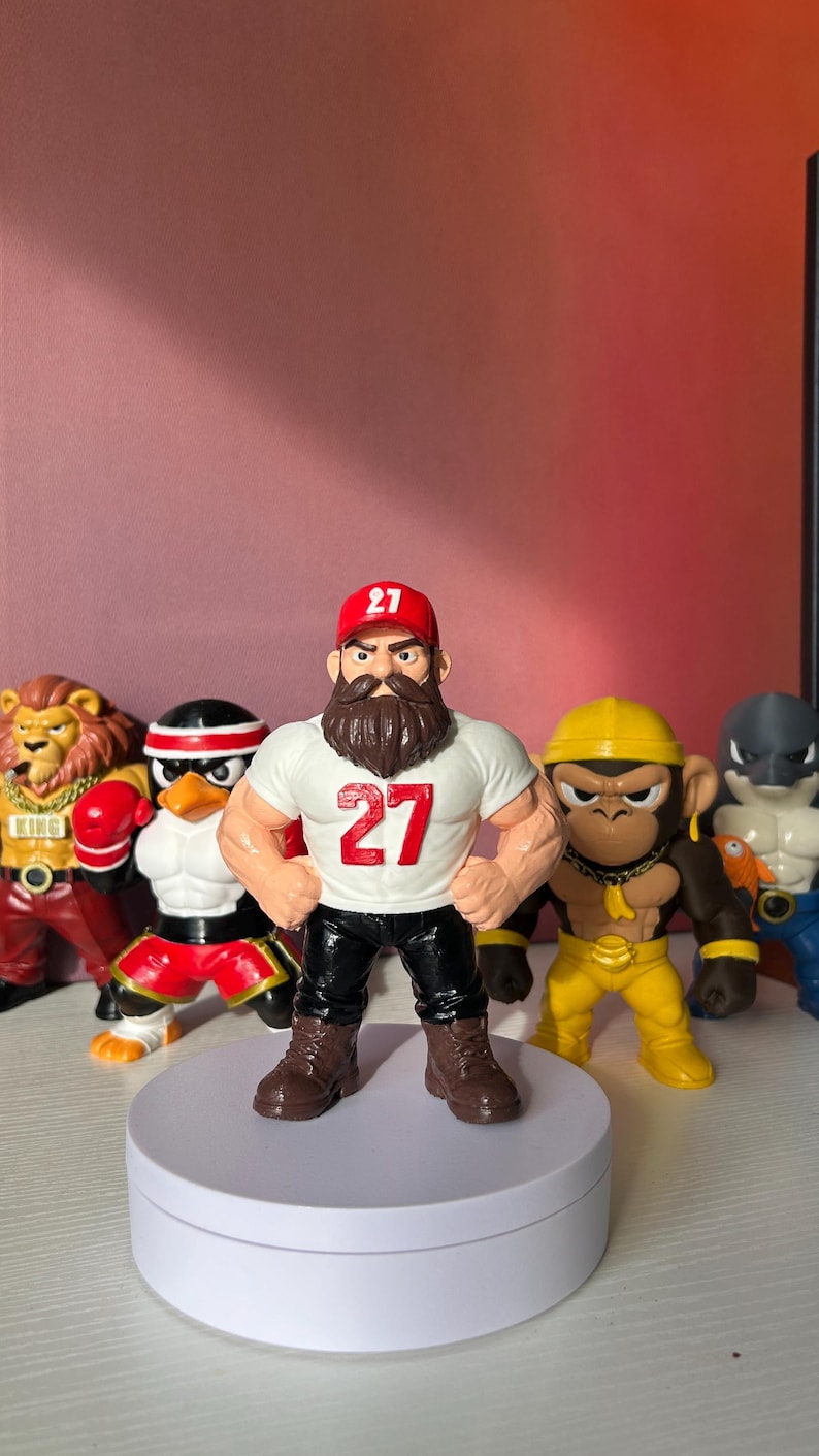 May include: A group of colourful figurines, including a muscular figure wearing a white t-shirt with the number "27" and a red cap. Other figures include a lion, penguin, monkey, and shark. The figures are displayed on a white rotating platform.