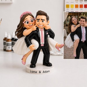 May include: A wedding cake topper depicting a bride and groom. The bride, in a white dress and pink heels, playfully pulls the groom's cheeks. The groom wears a black suit and pink tie. The base is inscribed with "Luna & Adam."