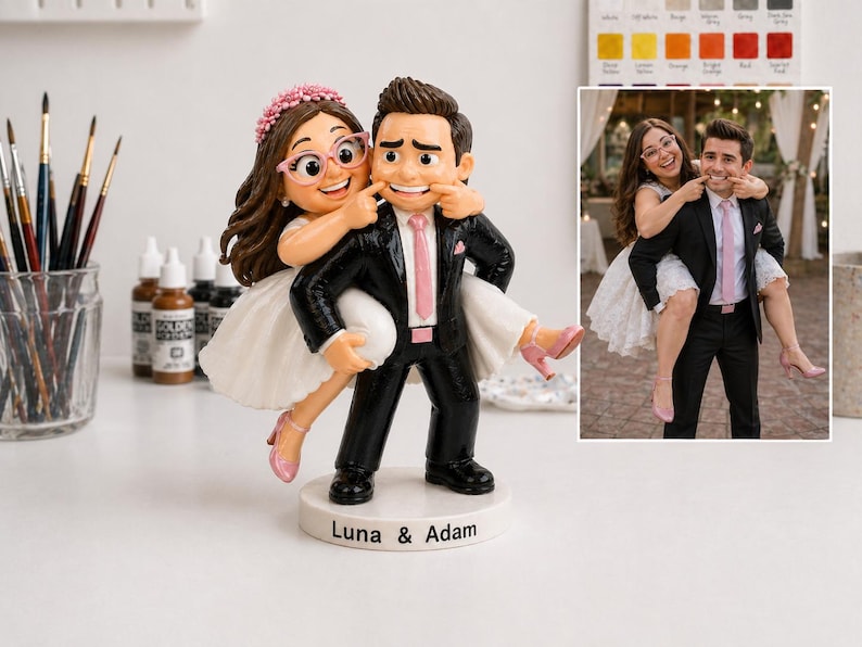 May include: A wedding cake topper depicting a bride and groom. The bride, in a white dress and pink heels, playfully pulls the groom's cheeks. The groom wears a black suit and pink tie. The base is inscribed with "Luna & Adam."