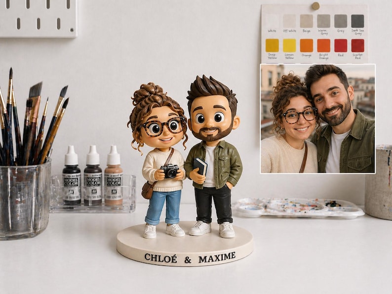 May include: A custom figurine of a couple, with the names "CHLOÉ & MAXIME" on the base. The figures are wearing glasses and casual clothing. The woman holds a camera, and the man holds a book. A photo of the couple is in the background.